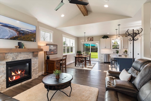 living room featuring dark wood-type flooring, ceiling fan with notable chandelier, a stone fireplace, and lofted ceiling with beams