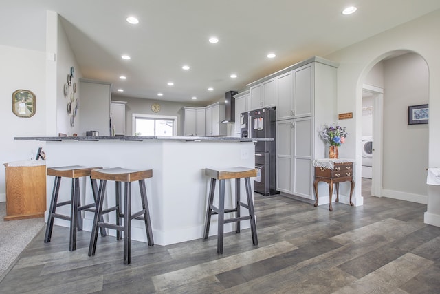 kitchen with a breakfast bar area, wall chimney exhaust hood, high end refrigerator, washer / clothes dryer, and dark stone countertops