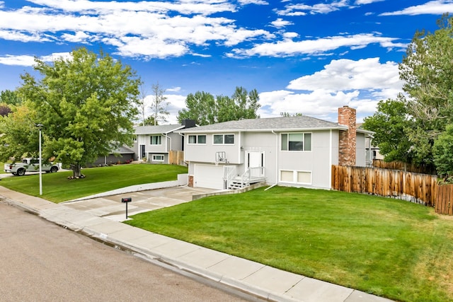 raised ranch featuring concrete driveway, a chimney, an attached garage, fence, and a front lawn