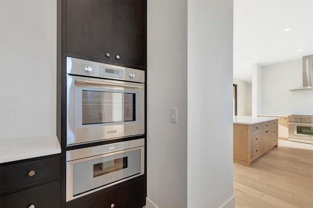 kitchen featuring double oven, stove, wall chimney exhaust hood, and light hardwood / wood-style flooring