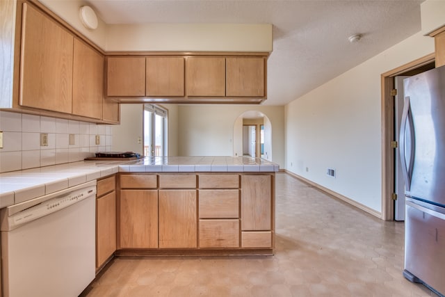 kitchen with kitchen peninsula, light brown cabinets, tile countertops, white dishwasher, and stainless steel fridge