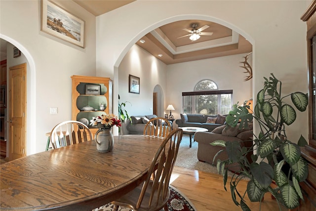 dining area with ceiling fan, arched walkways, wood finished floors, a towering ceiling, and a tray ceiling