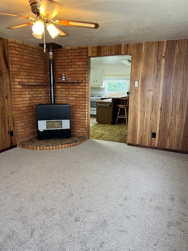 unfurnished living room featuring carpet flooring, a wood stove, wood walls, ceiling fan, and a textured ceiling