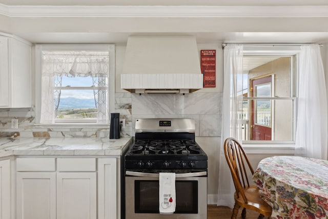 kitchen with custom exhaust hood, stainless steel gas stove, backsplash, white cabinetry, and light stone countertops