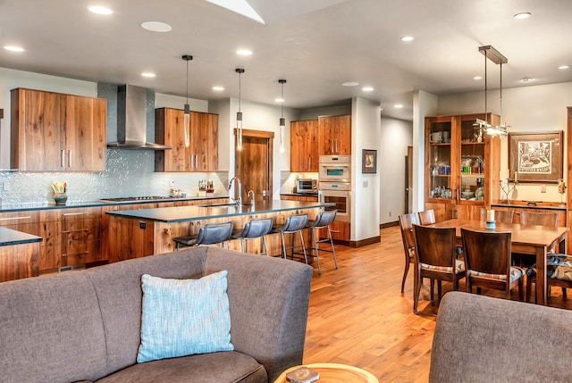 kitchen featuring brown cabinetry, dark countertops, a sink, a breakfast bar, and wall chimney exhaust hood