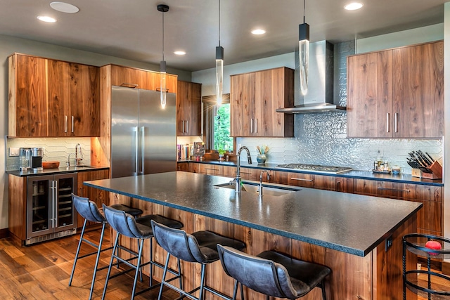 kitchen with wall chimney range hood, dark countertops, a sink, beverage cooler, and stainless steel appliances