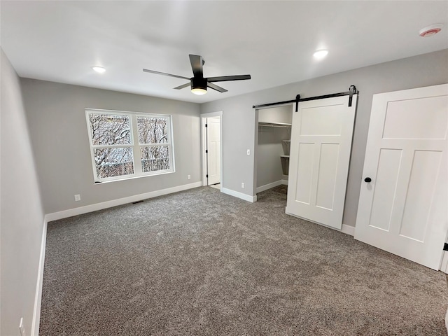 unfurnished bedroom featuring a walk in closet, a barn door, ceiling fan, and dark colored carpet