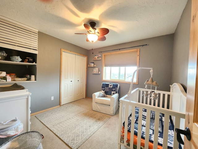 bedroom with a nursery area, a closet, a textured ceiling, ceiling fan, and light colored carpet