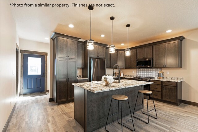 kitchen with light hardwood / wood-style floors, an island with sink, stainless steel appliances, decorative light fixtures, and light stone countertops