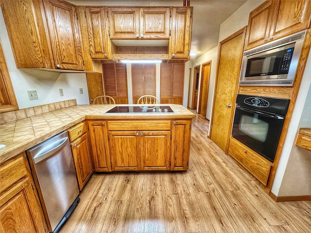 kitchen with brown cabinets, tile counters, light wood-type flooring, a peninsula, and black appliances