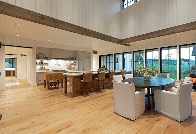 dining room featuring wooden walls, light hardwood / wood-style floors, beam ceiling, and a towering ceiling