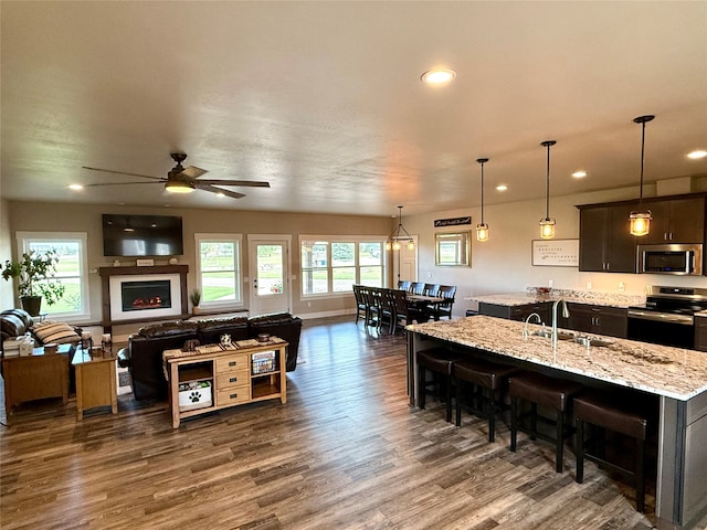 kitchen with light stone counters, an island with sink, decorative light fixtures, dark wood-type flooring, and appliances with stainless steel finishes