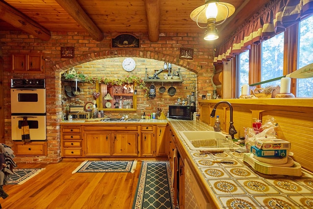 kitchen featuring beamed ceiling, wood-type flooring, double oven, brick wall, and tile countertops