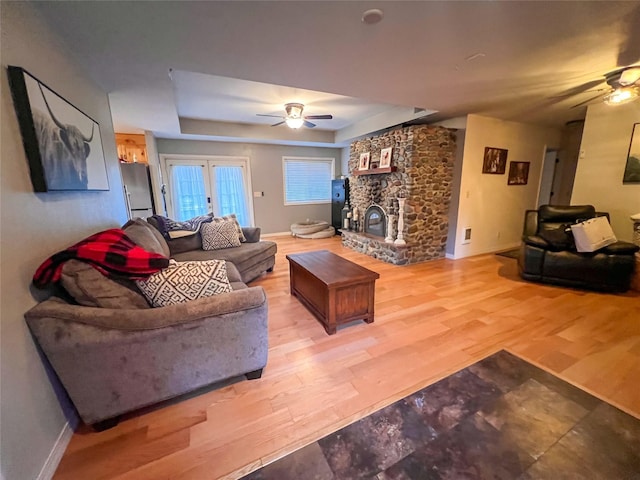 living room with a stone fireplace, a raised ceiling, ceiling fan, and light wood-type flooring