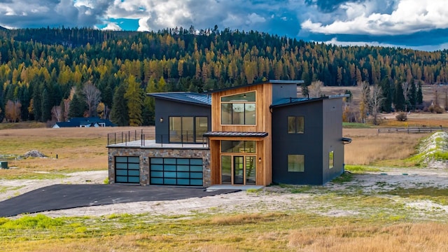 contemporary home with driveway, stone siding, metal roof, a standing seam roof, and a wooded view