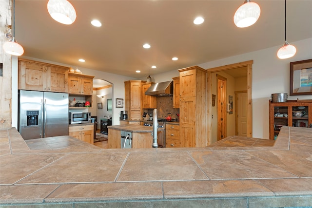 kitchen featuring light brown cabinetry, hanging light fixtures, stainless steel appliances, wall chimney exhaust hood, and decorative backsplash