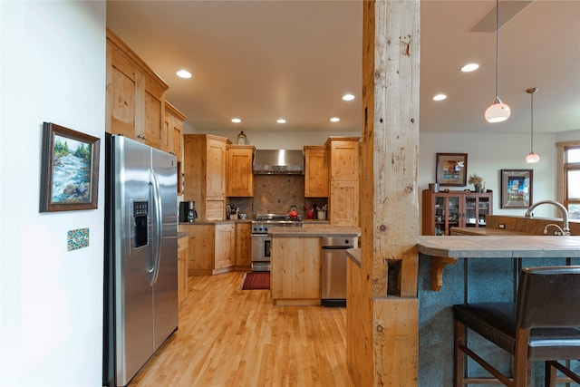 kitchen featuring appliances with stainless steel finishes, light wood-type flooring, wall chimney exhaust hood, decorative light fixtures, and a breakfast bar area