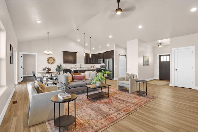 living room featuring light wood-type flooring, ceiling fan with notable chandelier, sink, a barn door, and high vaulted ceiling