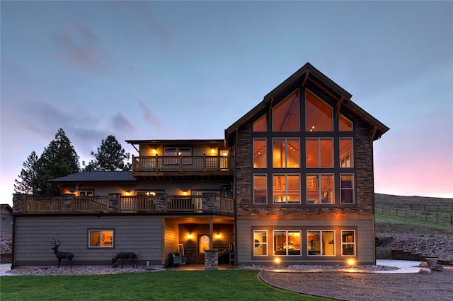 back house at dusk with a balcony, a patio area, and a lawn