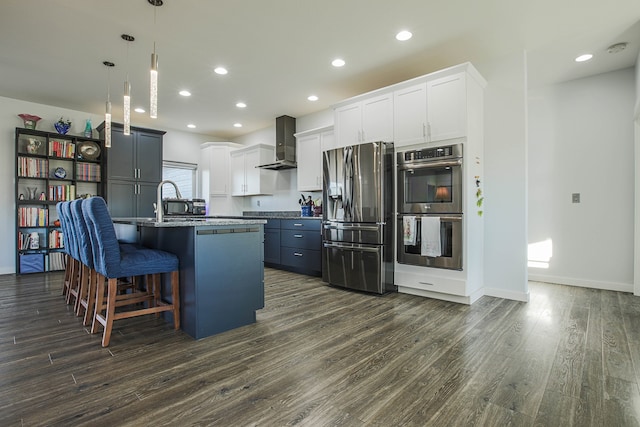 kitchen with wall chimney range hood, dark hardwood / wood-style flooring, appliances with stainless steel finishes, a breakfast bar, and a kitchen island with sink