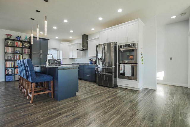 kitchen featuring appliances with stainless steel finishes, wall chimney exhaust hood, dark wood-style flooring, a kitchen island with sink, and a kitchen breakfast bar