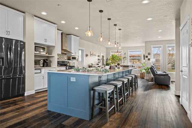 kitchen with wall chimney range hood, an island with sink, hanging light fixtures, white cabinetry, and stainless steel appliances