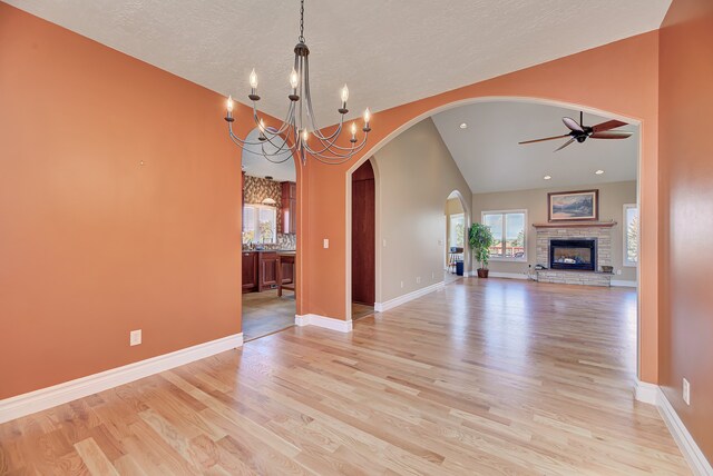 unfurnished dining area with a stone fireplace, vaulted ceiling, light wood-type flooring, a textured ceiling, and ceiling fan with notable chandelier