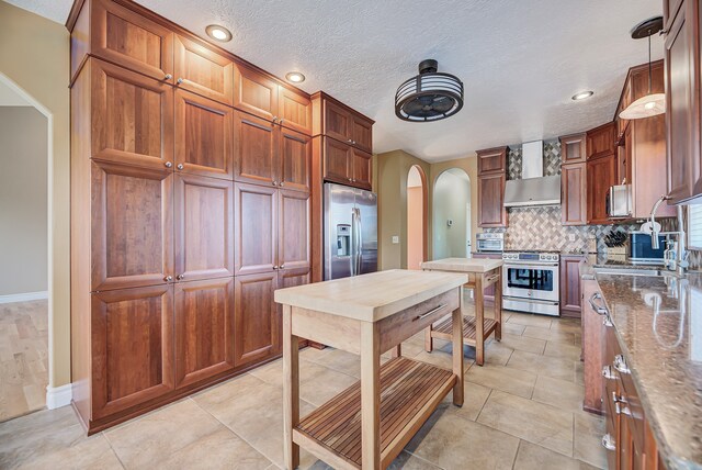 kitchen with stainless steel appliances, wall chimney exhaust hood, pendant lighting, dark stone countertops, and decorative backsplash