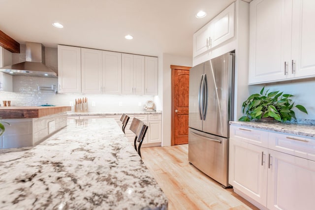 kitchen with wall chimney range hood, stainless steel fridge, light hardwood / wood-style flooring, light stone counters, and white cabinets