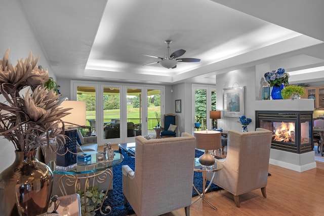 living room with ceiling fan, a tray ceiling, hardwood / wood-style floors, a multi sided fireplace, and french doors