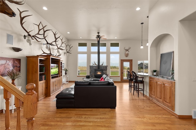 living room with a towering ceiling, ceiling fan, a wood stove, and light hardwood / wood-style floors