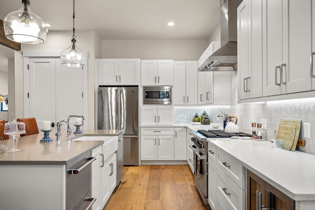 kitchen featuring white cabinetry, appliances with stainless steel finishes, wall chimney exhaust hood, hanging light fixtures, and light wood-type flooring