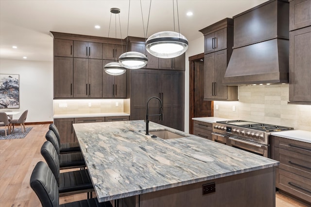 kitchen with light wood-type flooring, premium range hood, backsplash, stainless steel range, and a kitchen island with sink