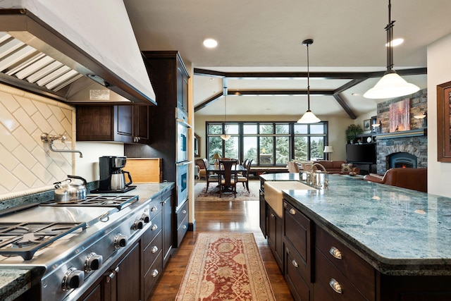 kitchen with wall chimney exhaust hood, dark hardwood / wood-style flooring, lofted ceiling with beams, a kitchen island with sink, and appliances with stainless steel finishes