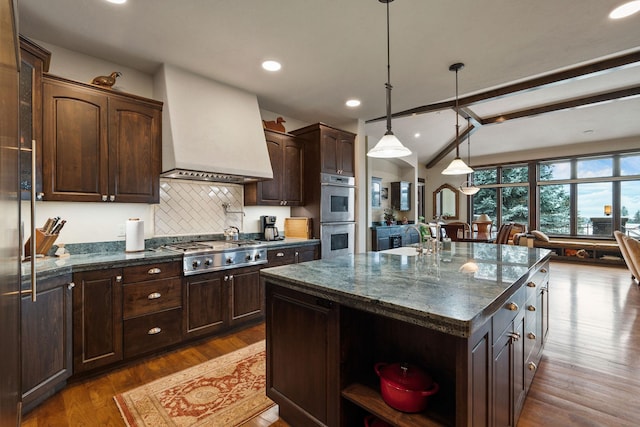 kitchen featuring custom exhaust hood, dark brown cabinets, a kitchen island with sink, and appliances with stainless steel finishes