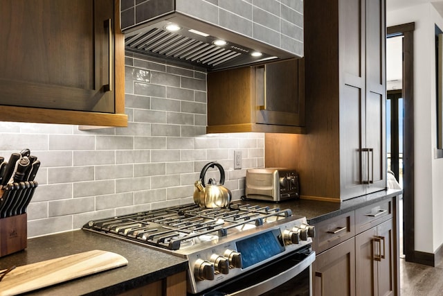 kitchen featuring wall chimney range hood, gas range, tasteful backsplash, dark brown cabinets, and baseboards