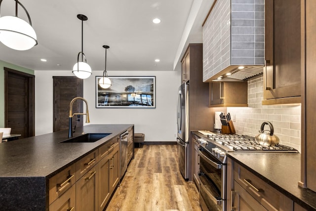 kitchen with appliances with stainless steel finishes, a kitchen island with sink, wall chimney range hood, sink, and hanging light fixtures