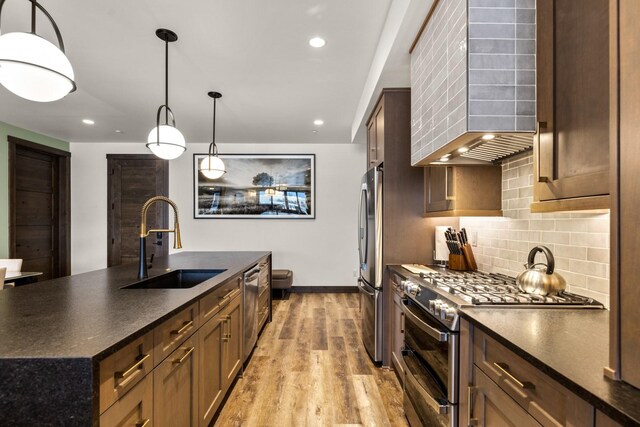 kitchen featuring appliances with stainless steel finishes, a sink, wall chimney range hood, light wood finished floors, and tasteful backsplash