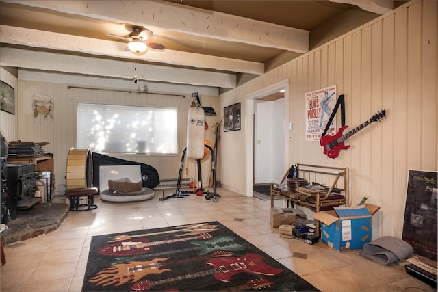 living room with ceiling fan, wooden walls, beamed ceiling, a wood stove, and light tile patterned flooring