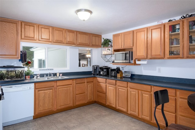 kitchen with glass insert cabinets, a sink, dark countertops, stainless steel microwave, and dishwasher