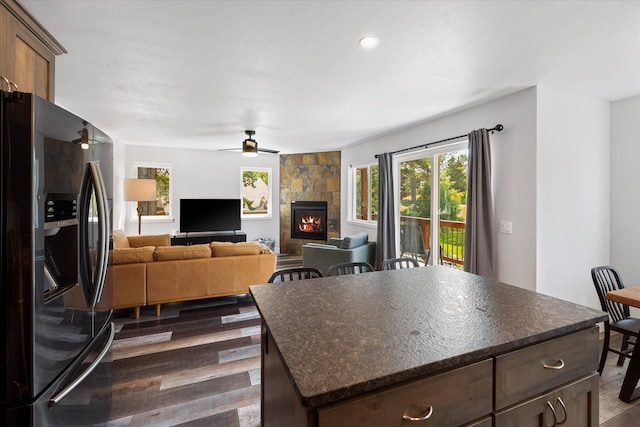 kitchen with a kitchen island, dark wood-type flooring, ceiling fan, a fireplace, and stainless steel refrigerator with ice dispenser