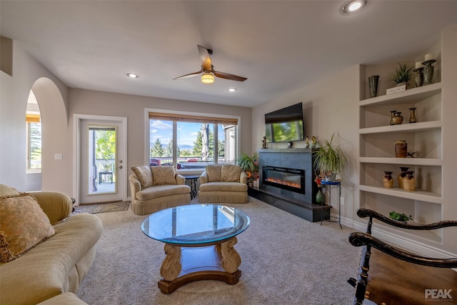 living room with a fireplace, built in shelves, light colored carpet, and ceiling fan