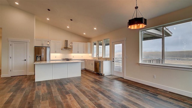 kitchen featuring wall chimney range hood, hanging light fixtures, appliances with stainless steel finishes, a kitchen island, and white cabinetry