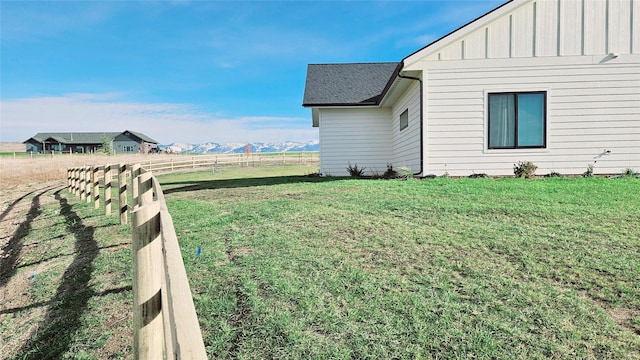 view of side of home with a rural view, a shingled roof, fence, a yard, and board and batten siding