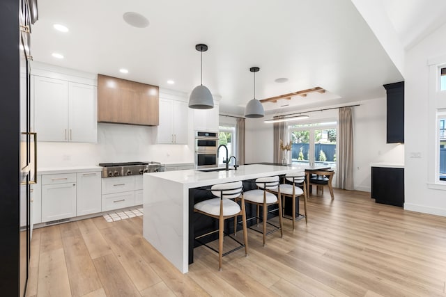 kitchen featuring stainless steel appliances, decorative light fixtures, a center island with sink, white cabinets, and light wood-type flooring