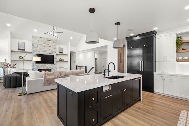 kitchen featuring a stone fireplace, sink, hanging light fixtures, light wood-type flooring, and an island with sink