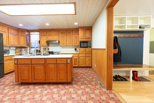 kitchen featuring sink, black microwave, wooden walls, and an island with sink