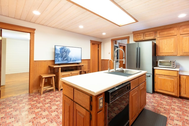 kitchen featuring stainless steel fridge, sink, a center island with sink, black dishwasher, and wood walls