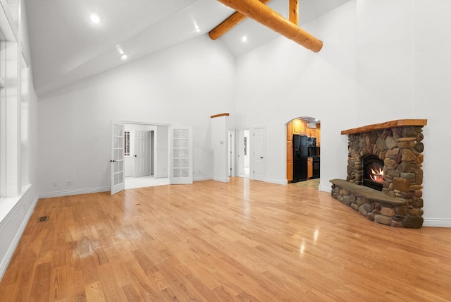 unfurnished living room featuring beamed ceiling, french doors, light hardwood / wood-style flooring, high vaulted ceiling, and a stone fireplace