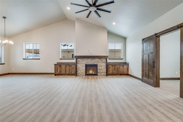 unfurnished living room with a stone fireplace, a barn door, light hardwood / wood-style floors, high vaulted ceiling, and ceiling fan with notable chandelier